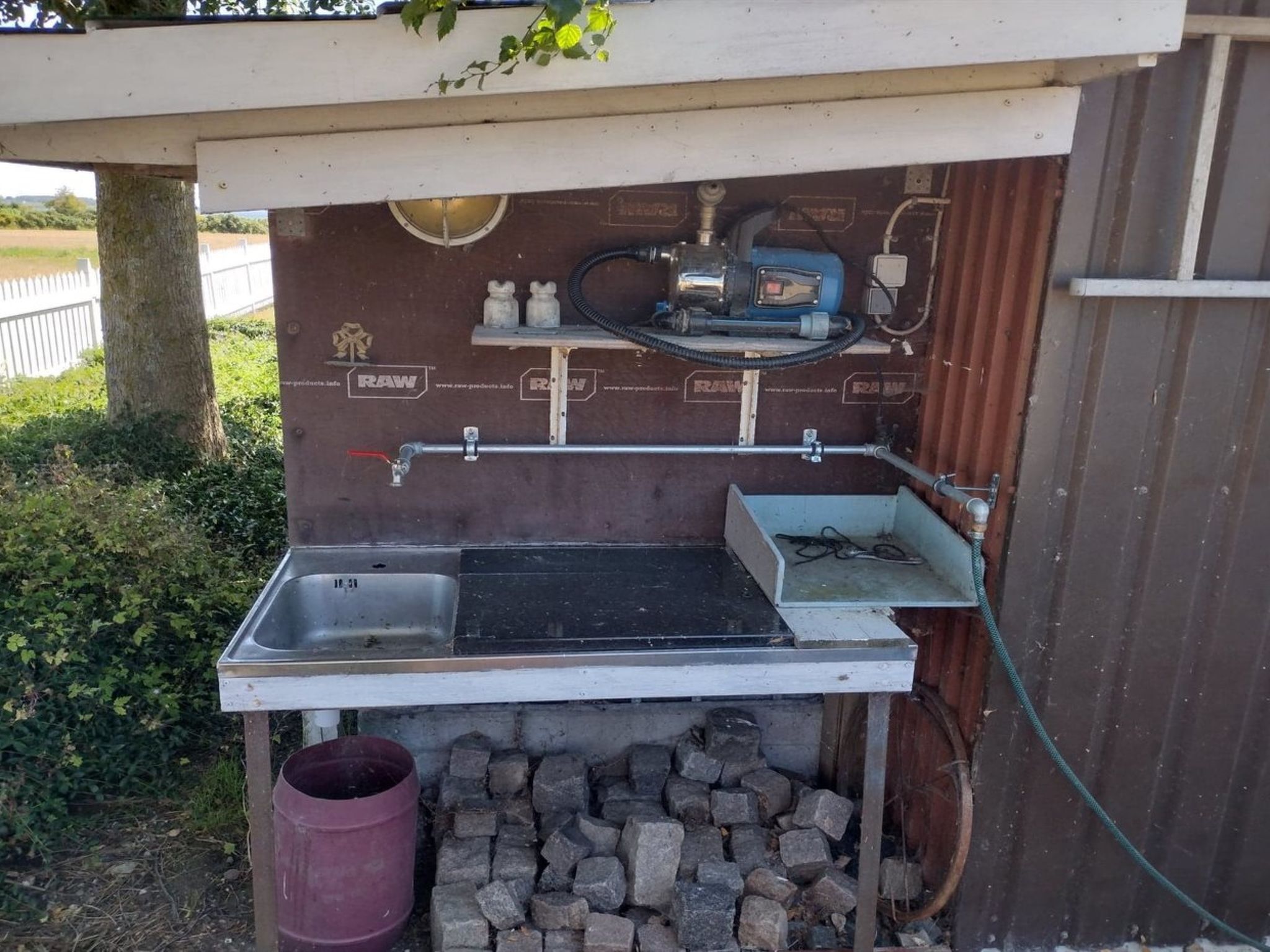 A small outdoor kitchen with a sink, a blue water pump, and a metal container.