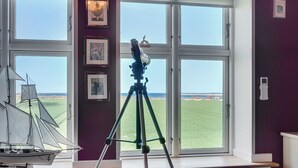 Ein Zimmer mit großen Fenstern, die auf ein grünes Feld blicken, einem auf einem Stativ stehenden Teleskop und gerahmten Bildern an der Wand.