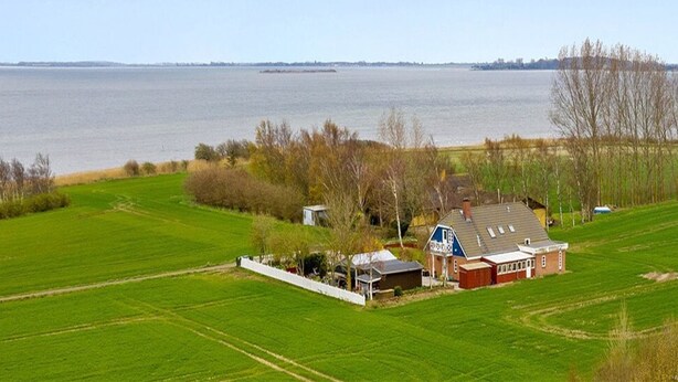 Ein ländliches Haus mit einem großen Feld im Vordergrund und einem Gewässer im Hintergrund.