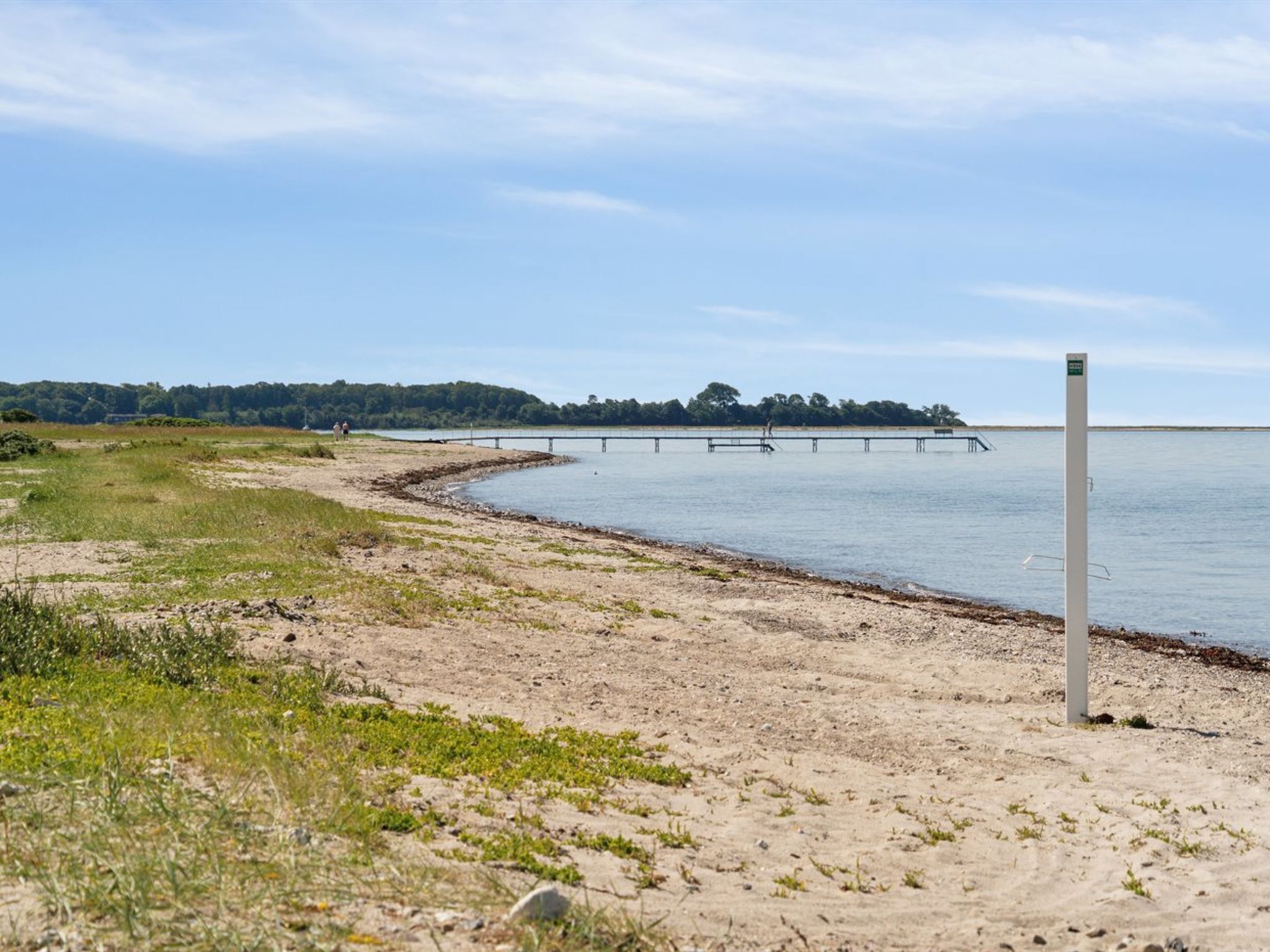 A sandy beach with a pier extending into the water, a grassy area, and a clear sky.