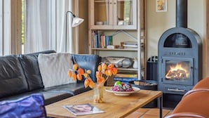Ein gemütliches Wohnzimmer mit einem Ledersofa, einem Couchtisch aus Holz mit einer Zeitschrift, einer Vase mit orangenen Blättern, einem Bücherregal und einem Kaminofen.