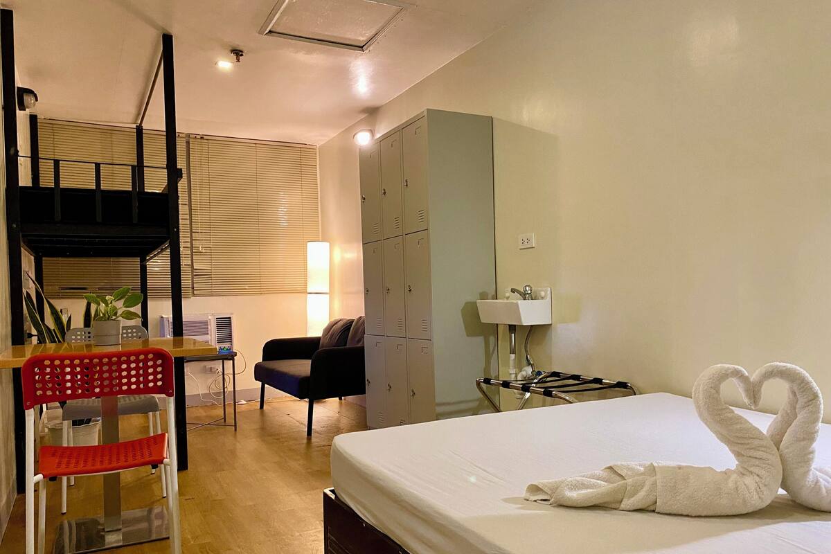 A hotel room with a bed, a desk with a chair, a wardrobe, a lamp, and a window with blinds.
