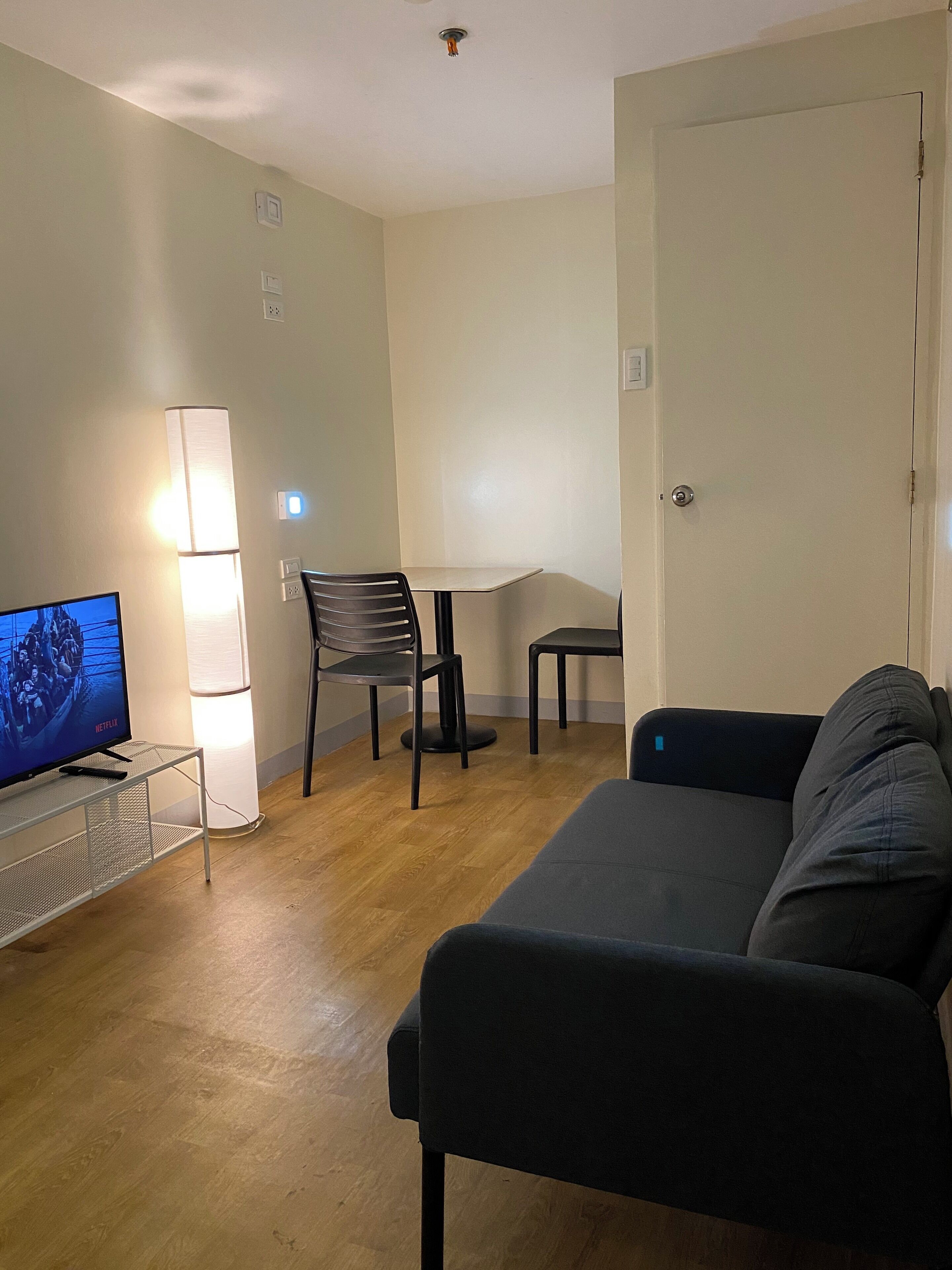 A small, furnished room with a sofa, a TV on a stand, a table, and a chair.