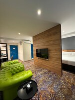 Habitación de hotel moderna con sofá verde, televisor de pantalla plana montado en una pared de madera y alfombra estampada.