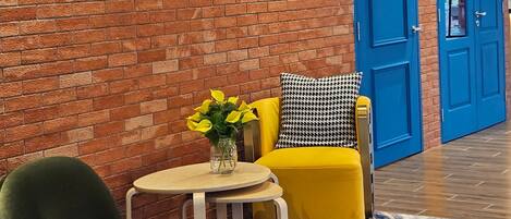 A lobby with a brick wall, a blue door, a yellow armchair, a green armchair, and a small table with a vase of yellow flowers.