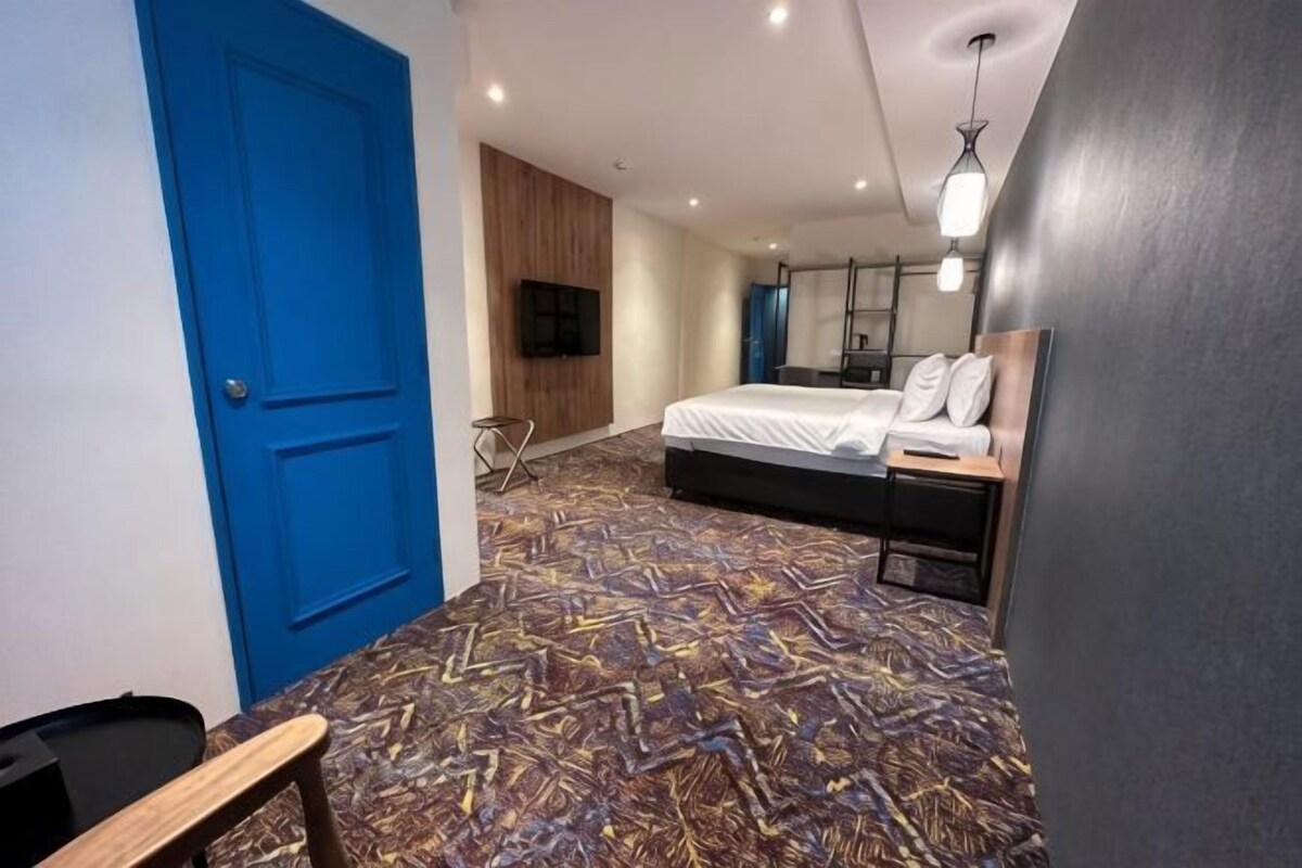 A hotel room with a patterned carpet, a bed, a desk, a chair, a blue door, and a television.