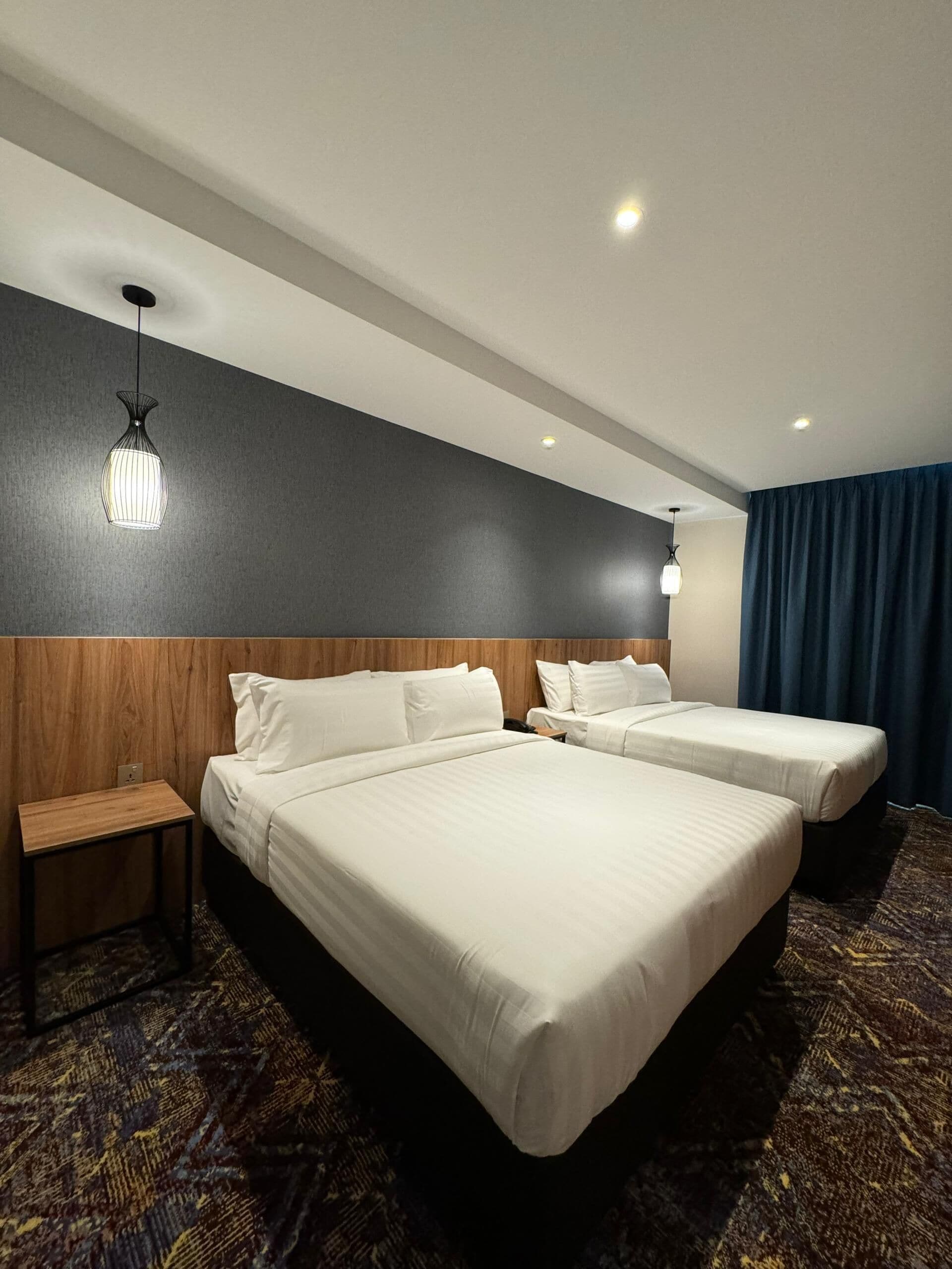 A hotel room with two beds, a wooden headboard, a bedside table, a patterned carpet, and a dark-colored curtain.