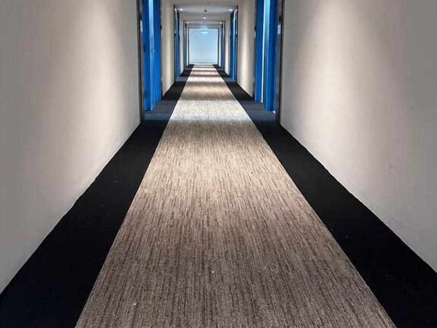Un long couloir étroit, doté d’un sol recouvert de moquette et de portes bleues de part et d’autre.
