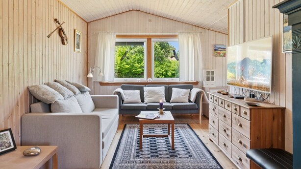 A cozy living room with a sofa, a coffee table, and a TV. There are windows with curtains, a wooden ceiling, and a view of greenery outside.