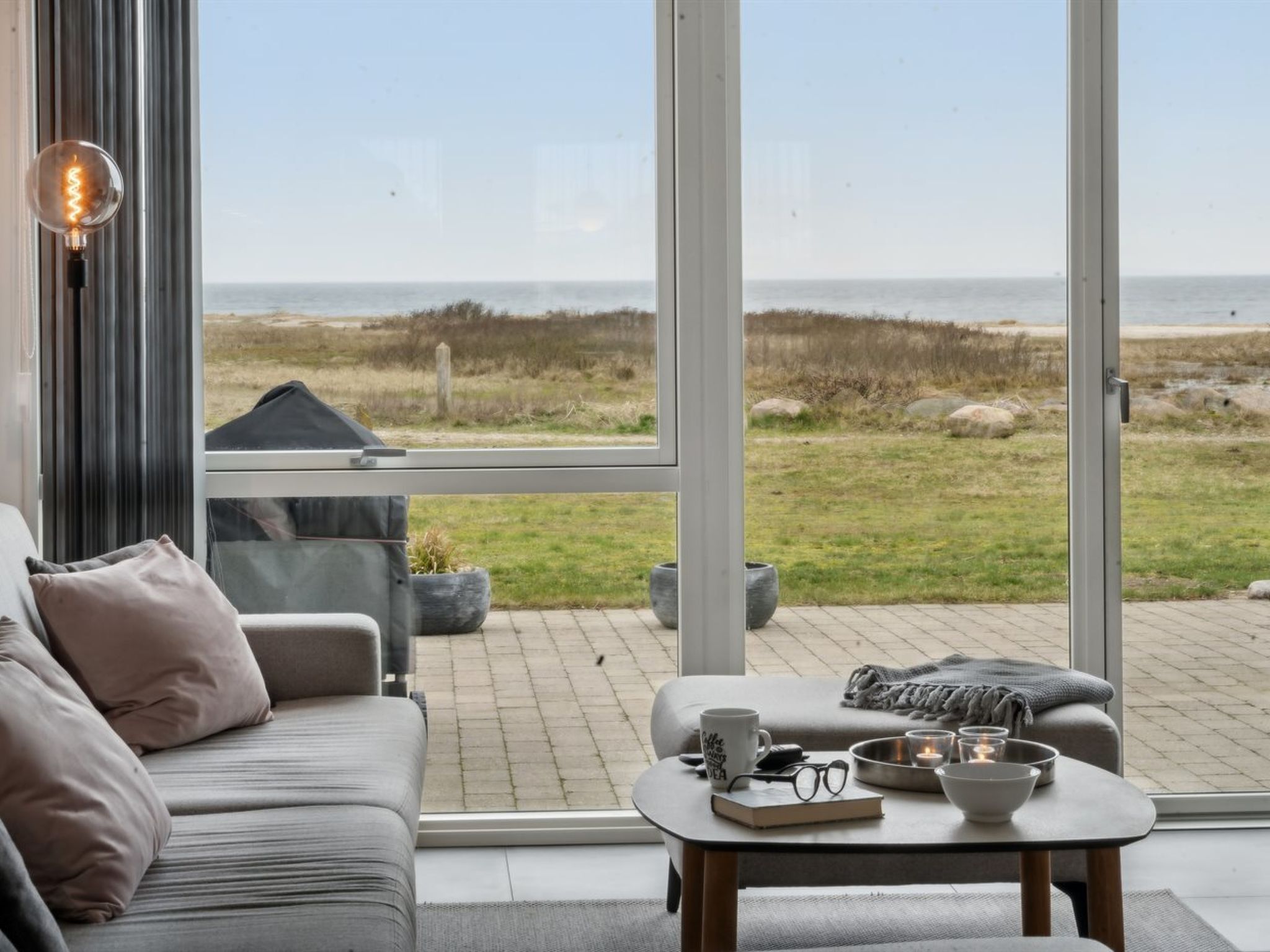 A modern living room with a large window overlooking a beach. There is a sofa, a coffee table with a book, glasses, and a mug.