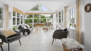 Une véranda spacieuse avec un haut plafond, de grandes fenêtres et un espace salon central comprenant un canapé et des fauteuils.