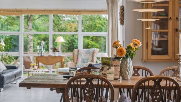 Un coin repas avec une table en bois, des chaises et un vase rempli de fleurs. Une fenêtre offre une vue sur la verdure à l’extérieur.