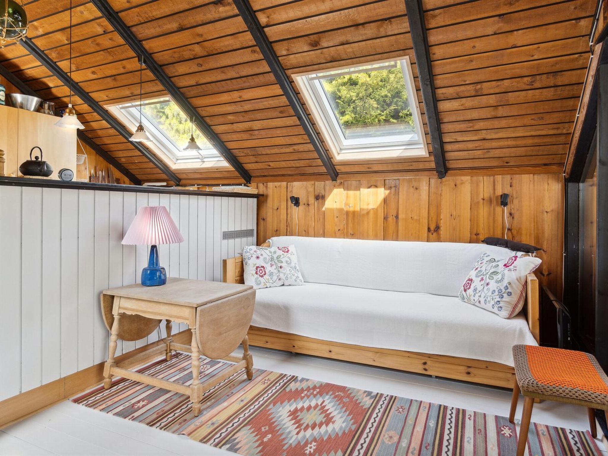 Een gezellig slaapkamer op zolder met een houten plafond, twee dakramen, een witte bank, een klein tafeltje en een kleurrijk vloerkleed.