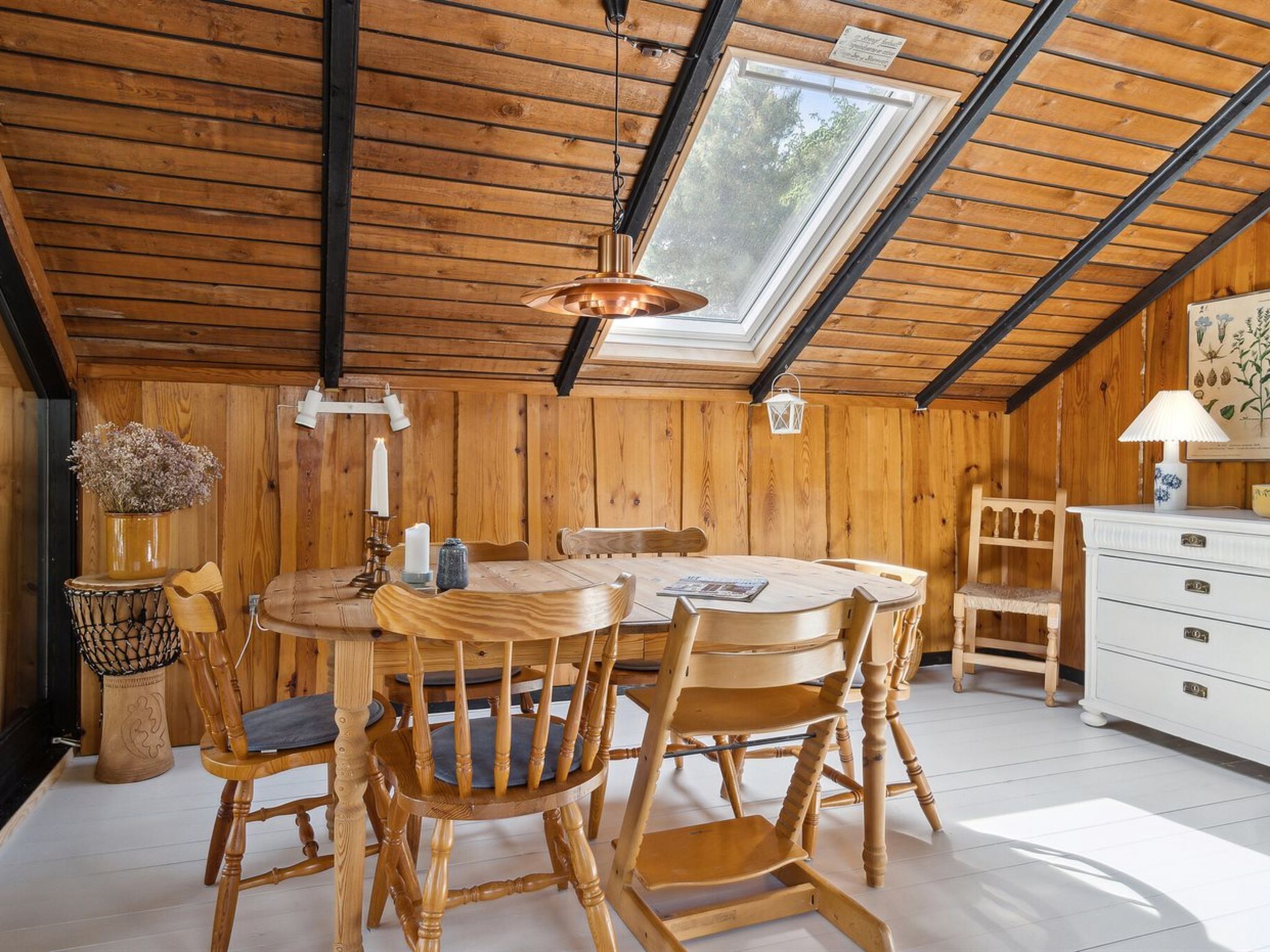 A cozy wooden room with a round table and chairs, a white dresser, and a skylight.