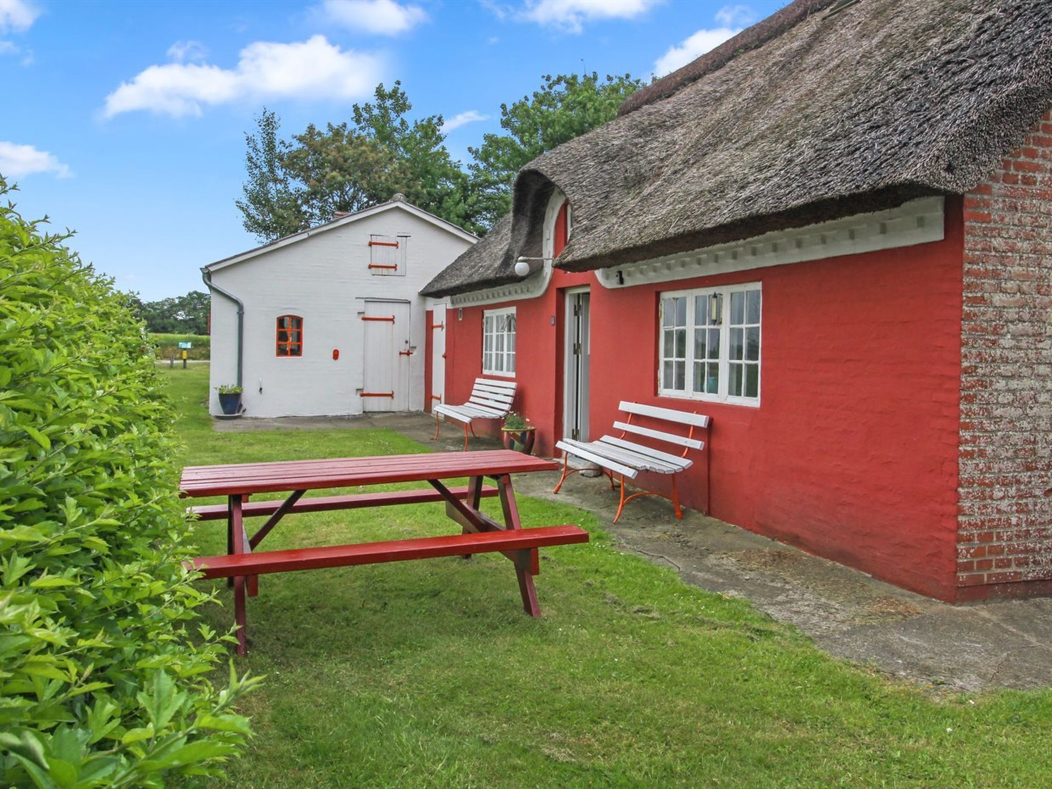 A red brick building with a thatched roof and a white building in the background. There is a picnic table and two benches in the foreground.