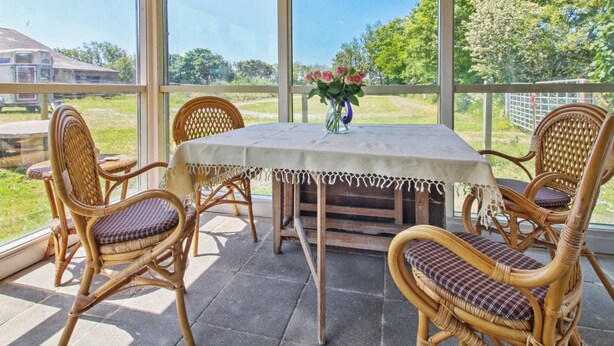 A covered patio with a table and wicker chairs, large windows offering a view of greenery and a distant building. - "Saarah" - 25km from the sea by Interhome (Bredebro)