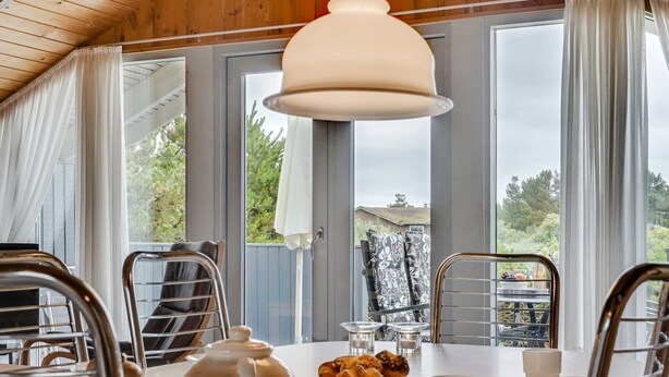 A dining area with a round table, metal chairs, a white teapot, and a view of the outdoors through large windows.