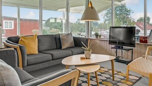 Un salon moderne avec un canapé gris, une table basse ronde et un téléviseur. De grandes fenêtres offrent une vue sur l’extérieur.