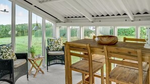 Un patio abrité avec une table et des chaises en bois, un fauteuil en osier avec un coussin, et une vue sur un paysage verdoyant à travers de grandes fenêtres.
