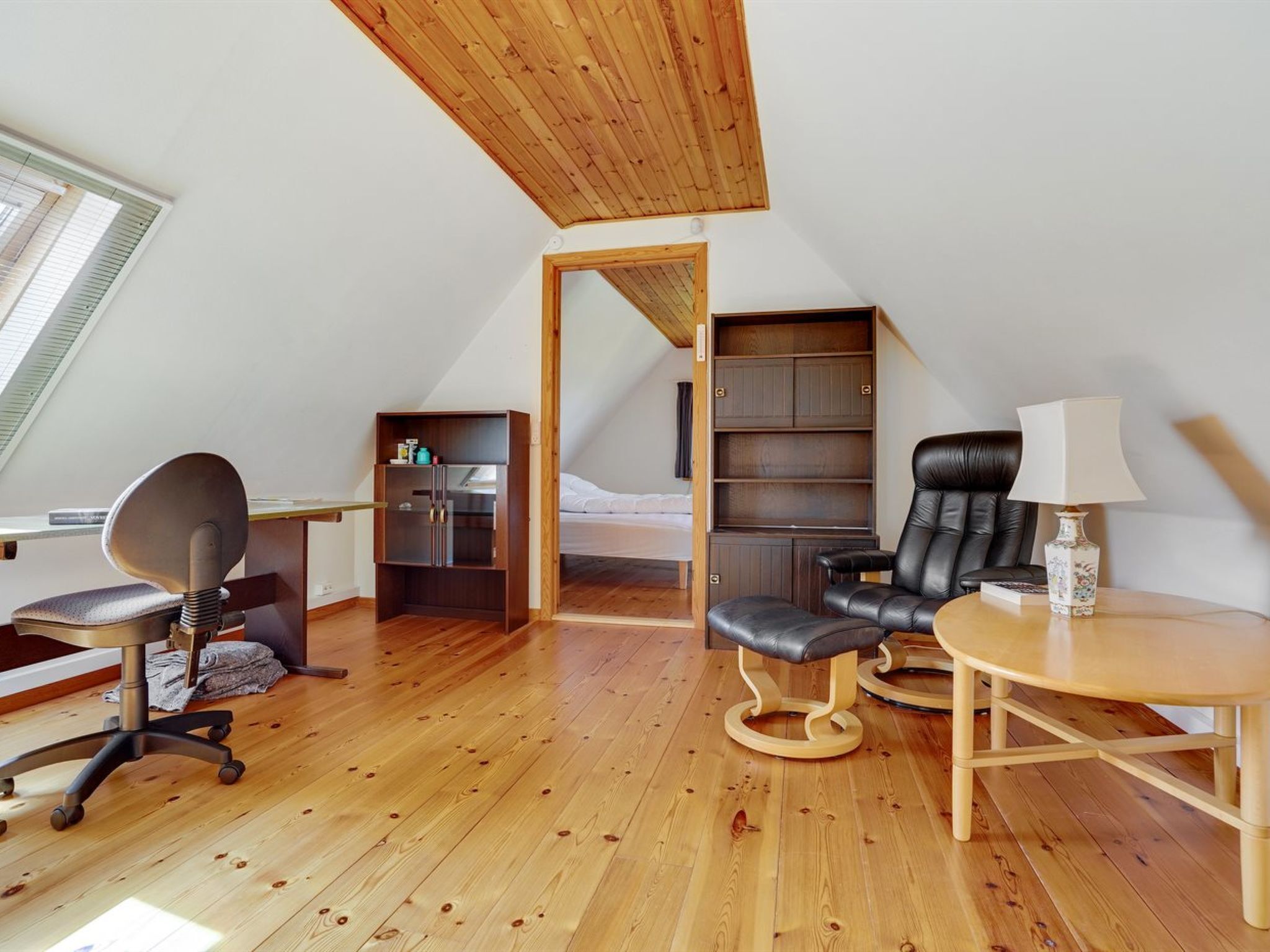 A loft bedroom with a sloped ceiling, wooden flooring, a desk with a chair, a bed, a bookshelf, and a small table with a lamp.