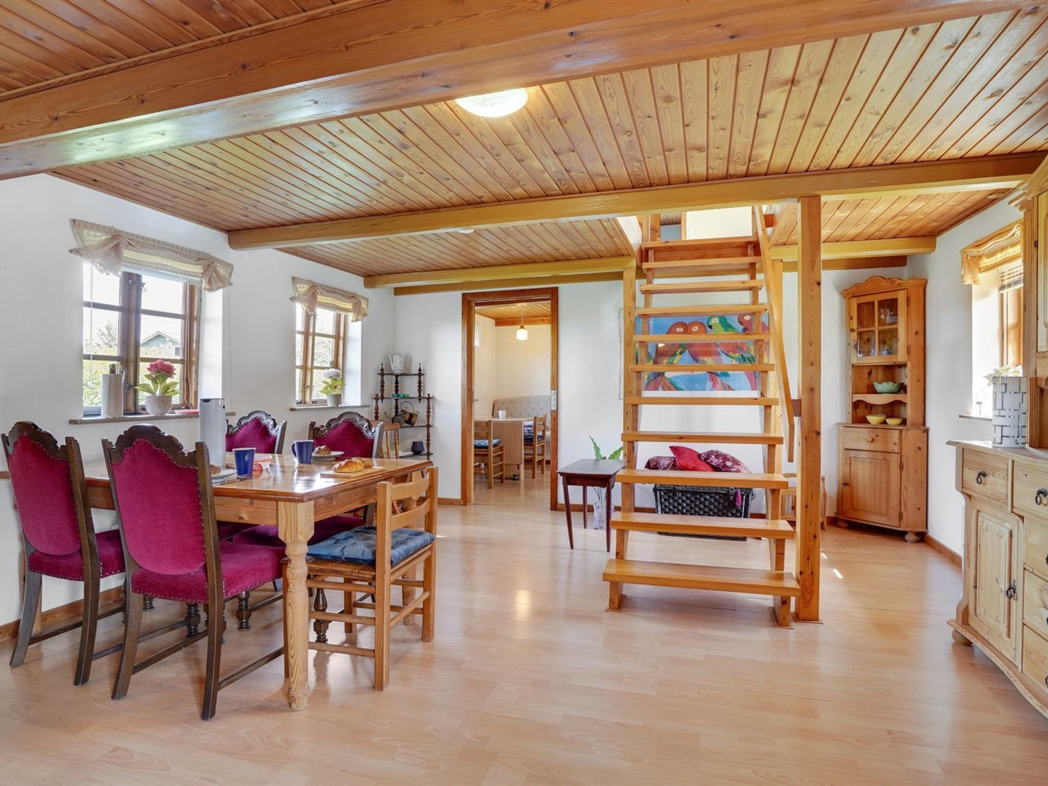 A spacious room with a wooden ceiling, a dining table with chairs, a ladder, and a kitchen area with wooden cabinets.