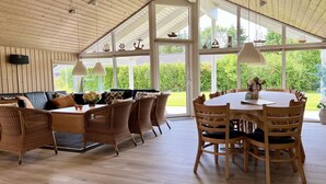 Una habitación amplia con techo alto, ventanas grandes, una zona de comedor con una mesa redonda y sillas, y un espacio de descanso con sofás y sillas de mimbre.