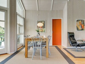 A dining area with a wooden table and chairs, a painting on the wall, and a bright orange door.