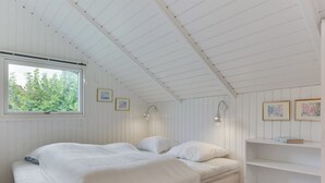 Ein Schlafzimmer mit Bett, Holzdielen, einem Fenster und einer weißen Wand mit Regalen und Bilderrahmen.