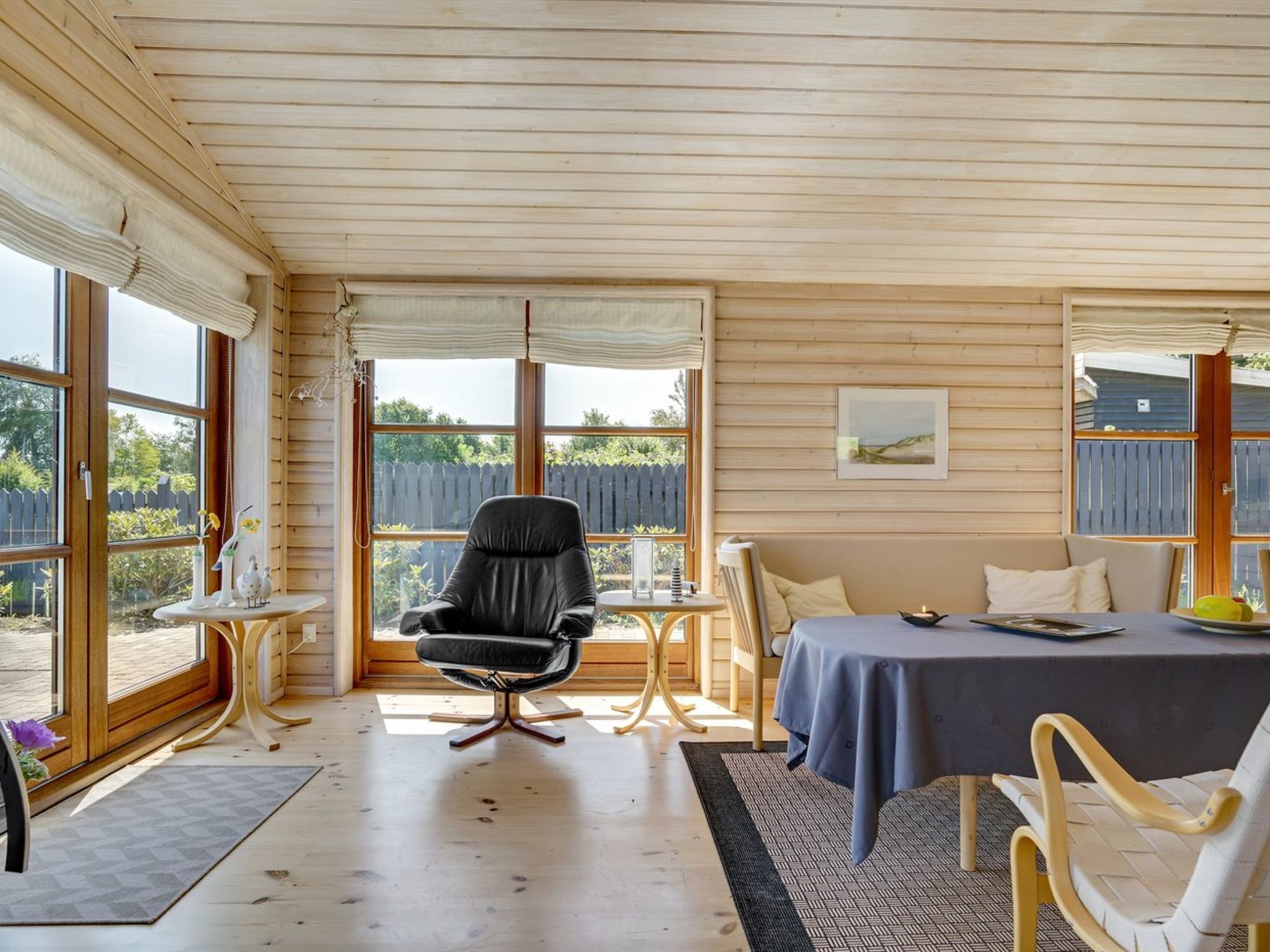 A cozy room with a wooden ceiling, large windows, a black chair, a dining table with a blue tablecloth, and a small side table.