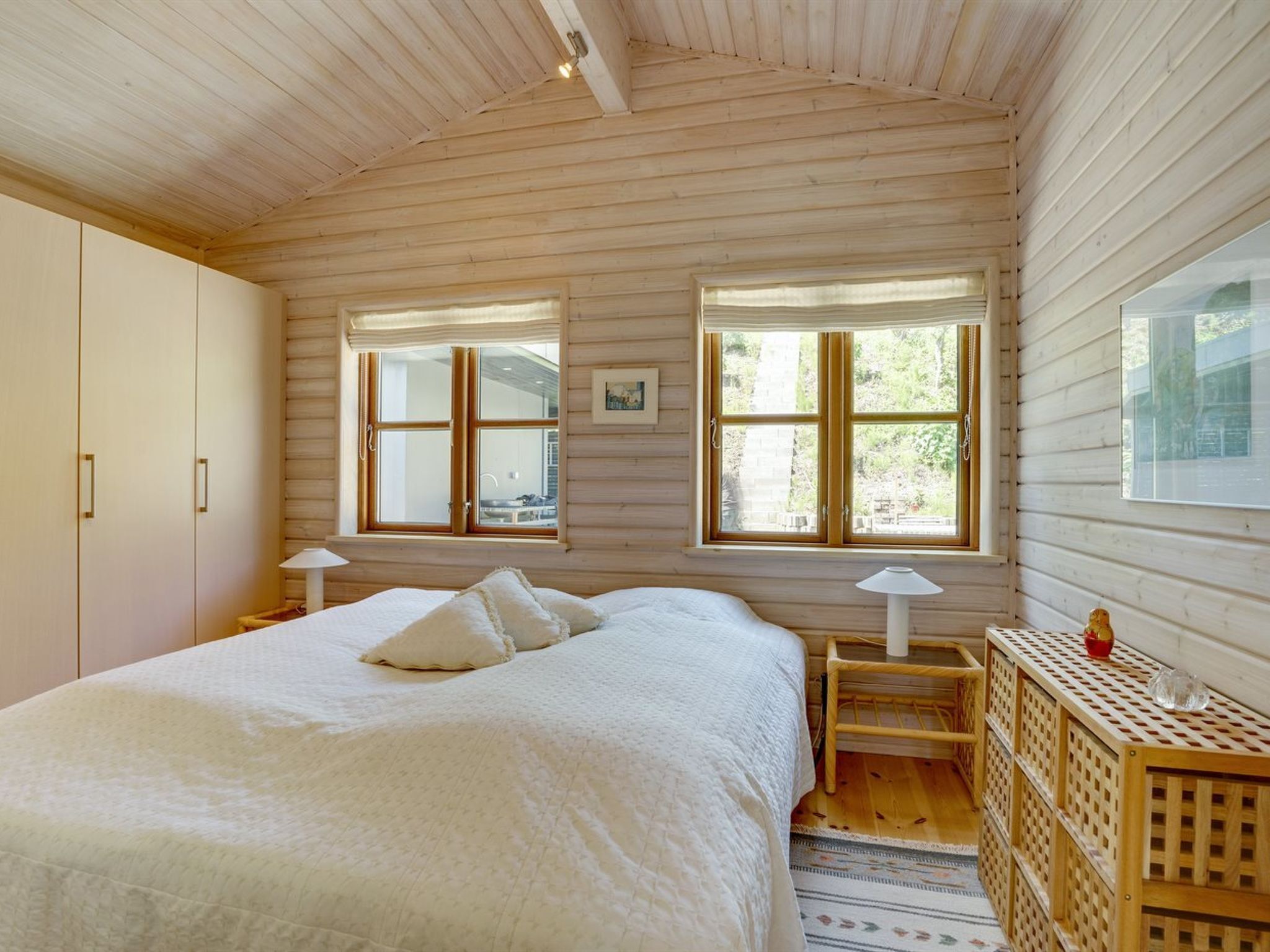 A bedroom with a large bed, two windows, a wooden wardrobe, and a small table with a lamp.
