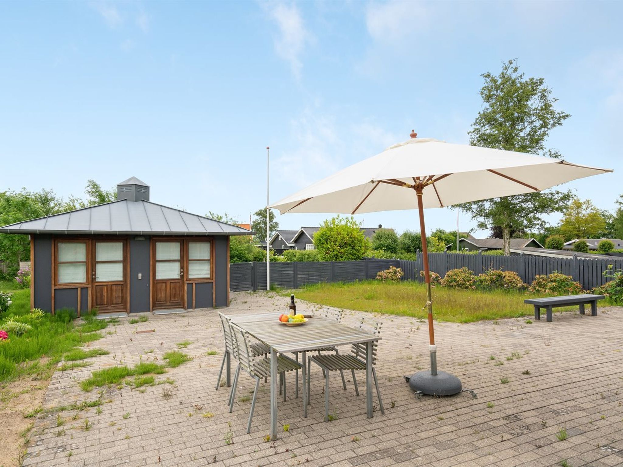 Une terrasse avec une table et des chaises, un grand parasol et un petit bâtiment doté d’un toit en pente.