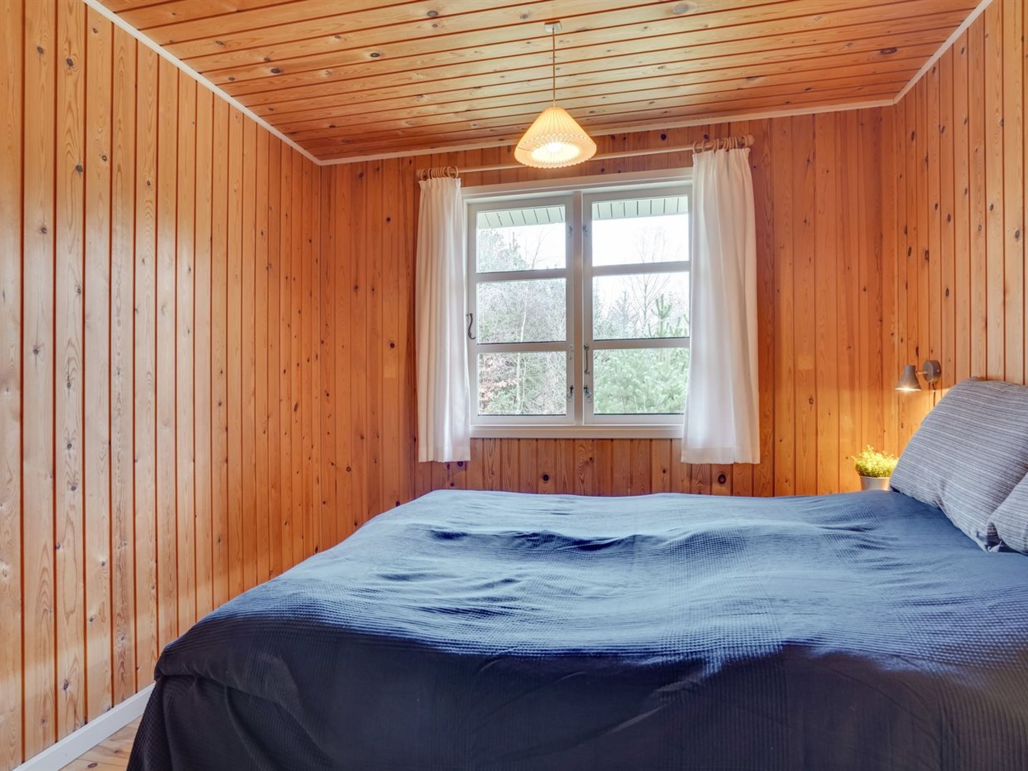 A wooden-paneled room with a bed, a window with curtains, and a hanging light fixture.