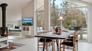 Ein modernes Interieur mit einem großen Esstisch, Holzstühlen, einem Kamin, einer Couch und einem großen Fenster mit Blick auf Bäume.
