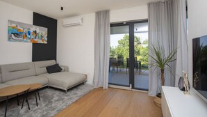 A modern living room with a grey sofa, a small wooden table, and a TV on a white stand. There is a large window with curtains, a painting on the wall, and an air conditioning unit. - Mondo Sole in Sveti Bartul (Rasa)