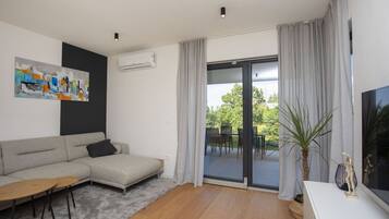 A modern living room with a grey sofa, a small wooden table, and a TV on a white stand. There is a large window with curtains, a painting on the wall, and an air conditioning unit.