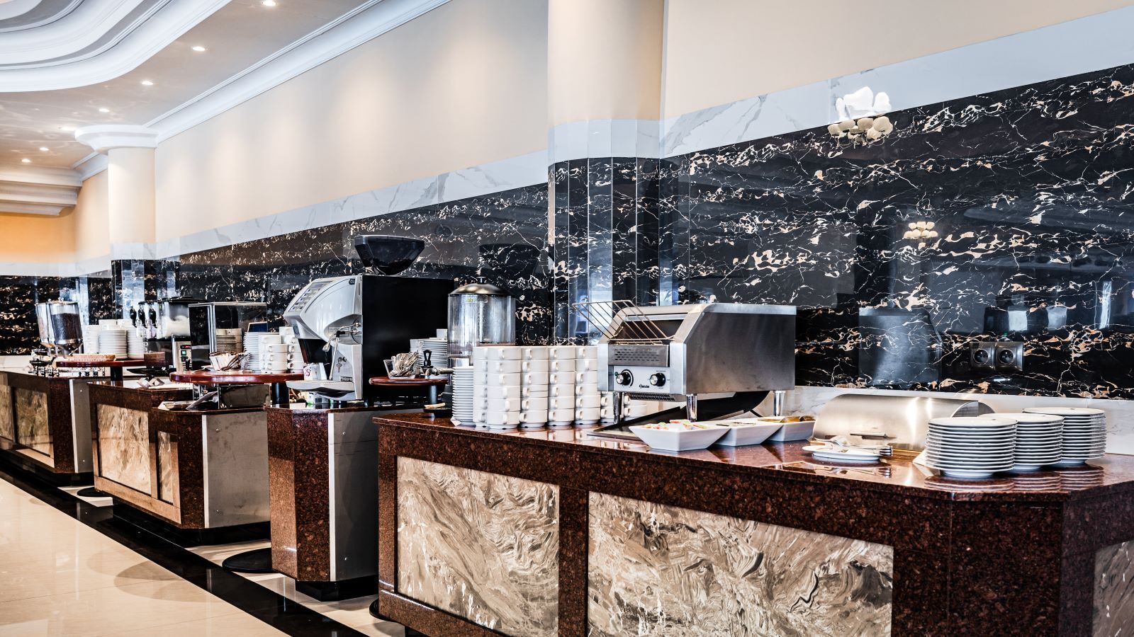 A modern hotel lobby with marble countertops, multiple coffee machines, and neatly stacked plates.