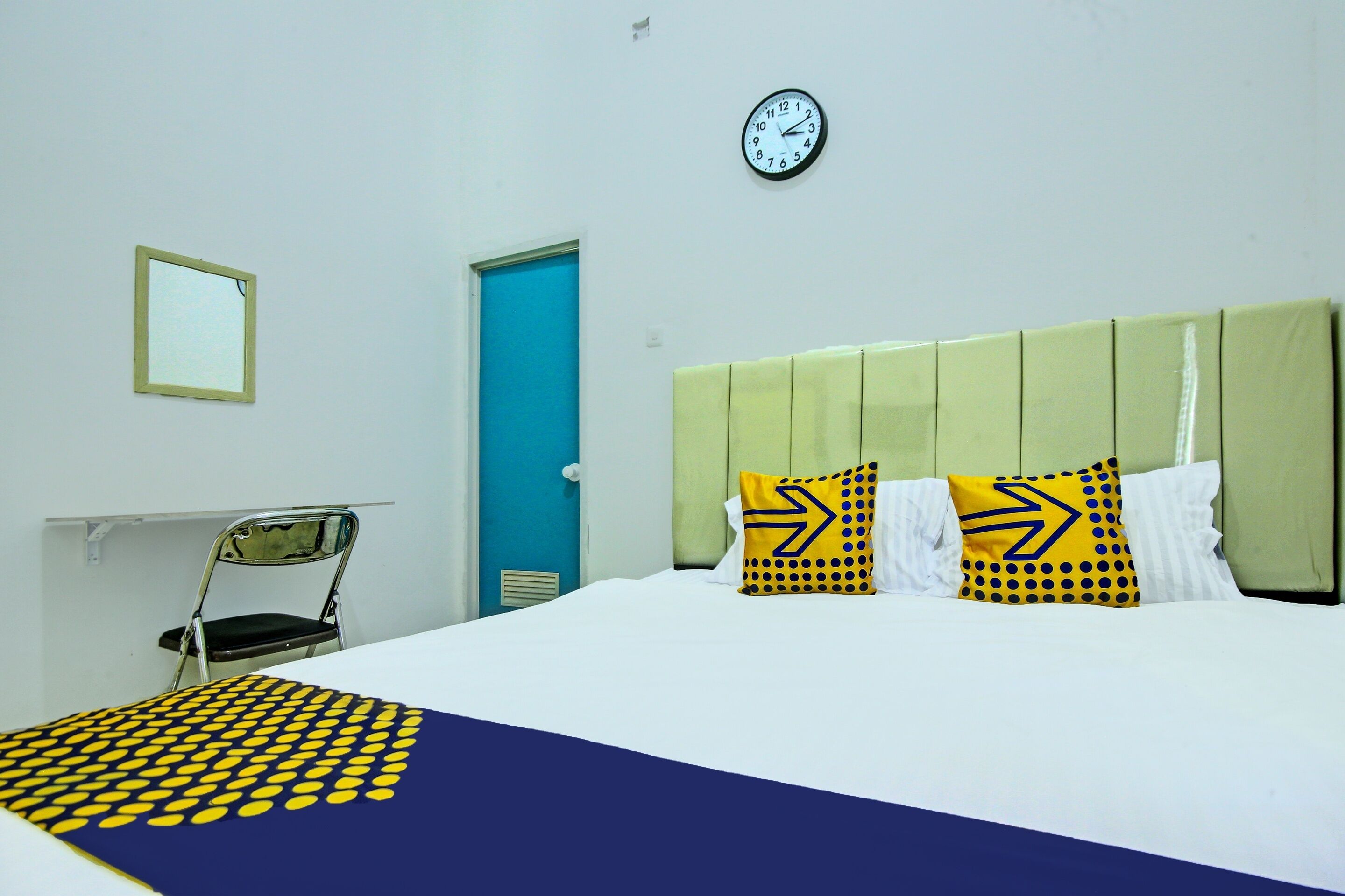 A hotel room with a bed, a chair, a clock, and a door.