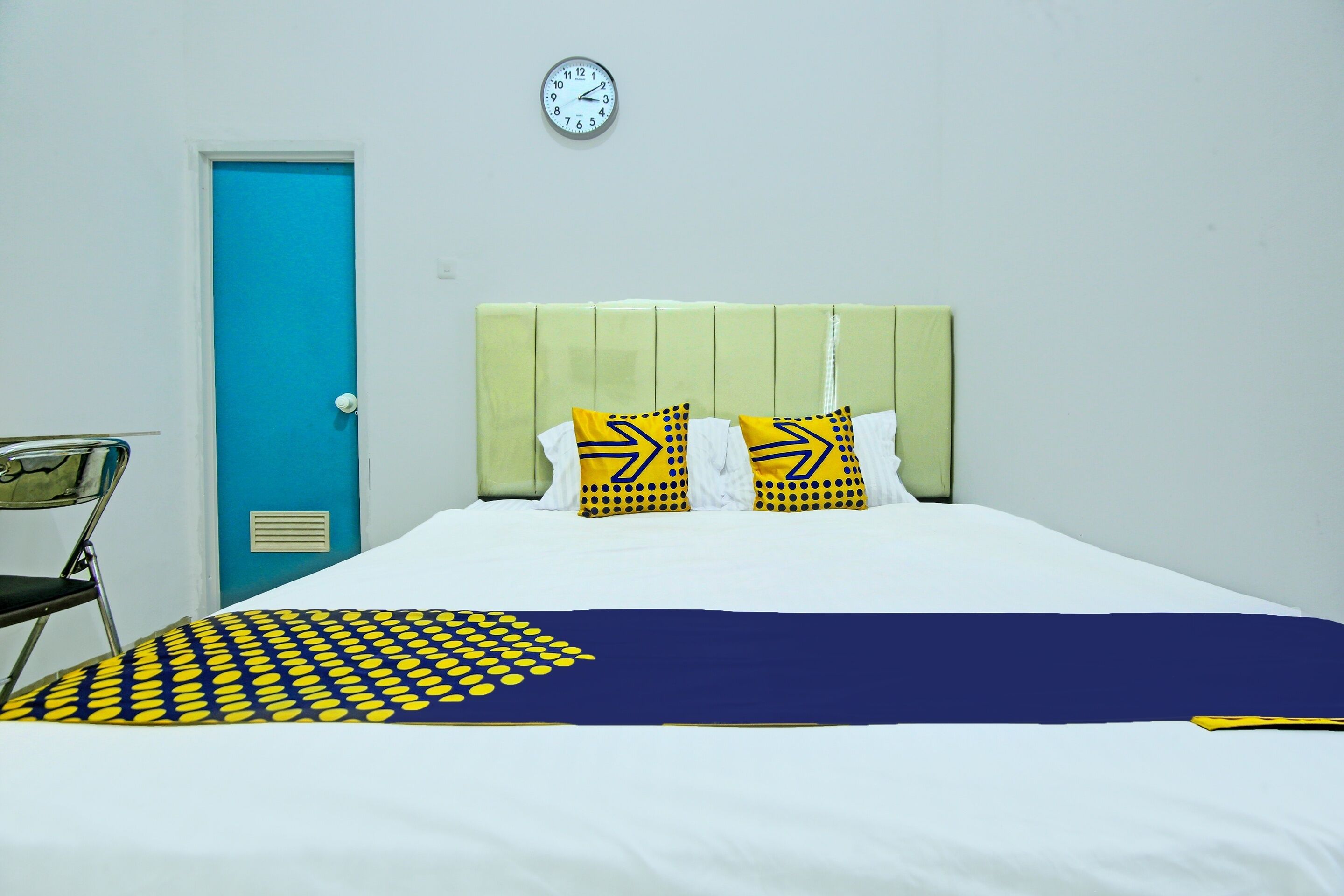 Ein Hotelzimmer mit einem Bett, einer blauen Tür, einer Wanduhr und einem Stuhl.