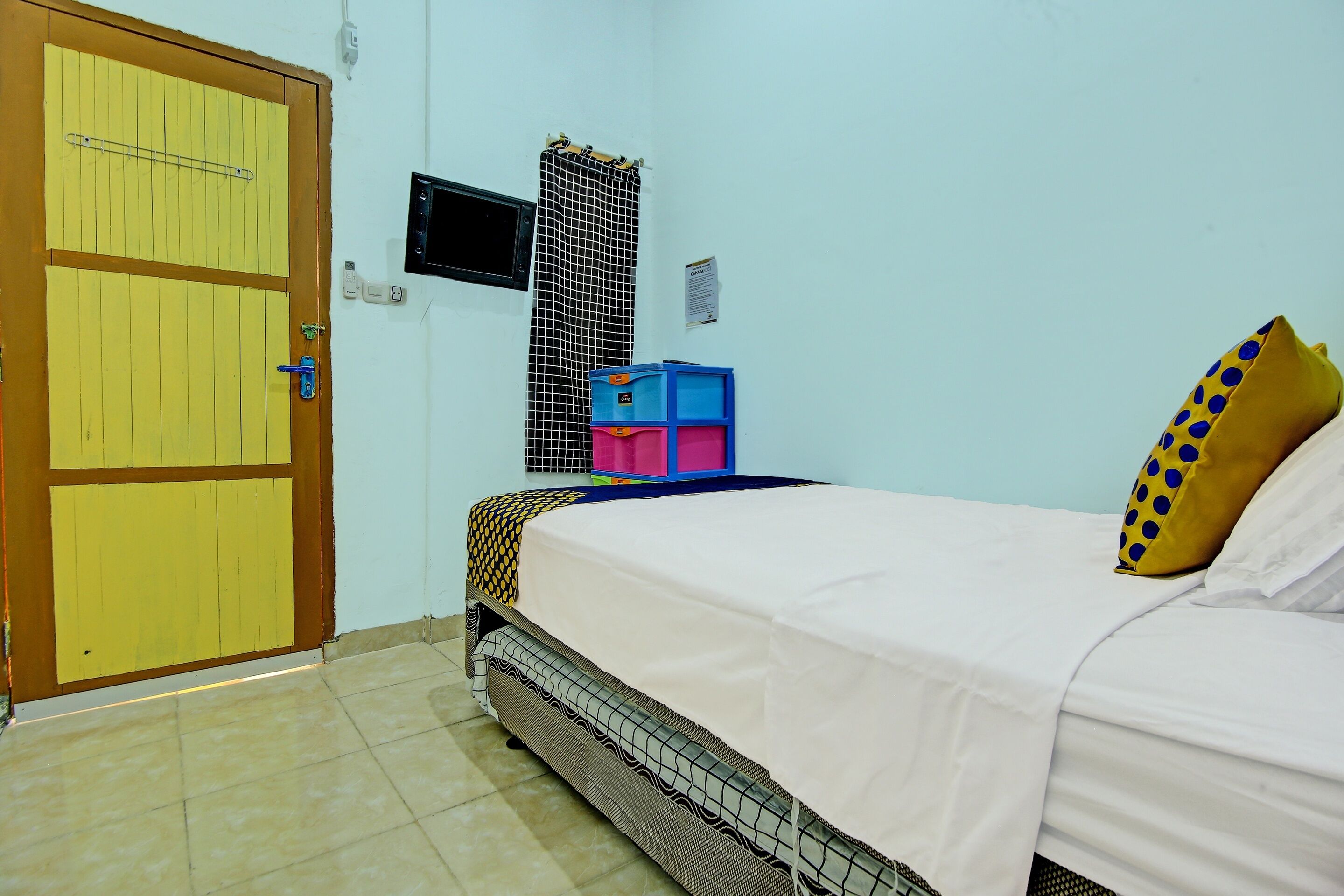 Una habitación con cama, puerta, televisor pequeño y ventana con mosquitero.