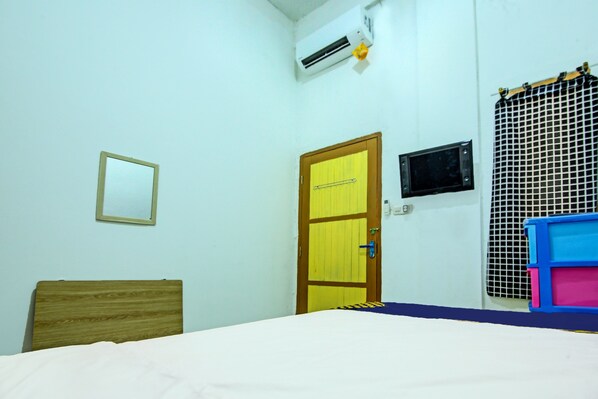 A room with a bed, a wooden headboard, a yellow door, an air conditioning unit, a small television, and a window with a metal grill.