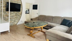 A modern living room with a stone wall, a round table with a marble top, a beige sofa, and a television mounted on the wall. - Apartment MARINA (Bremerhaven)