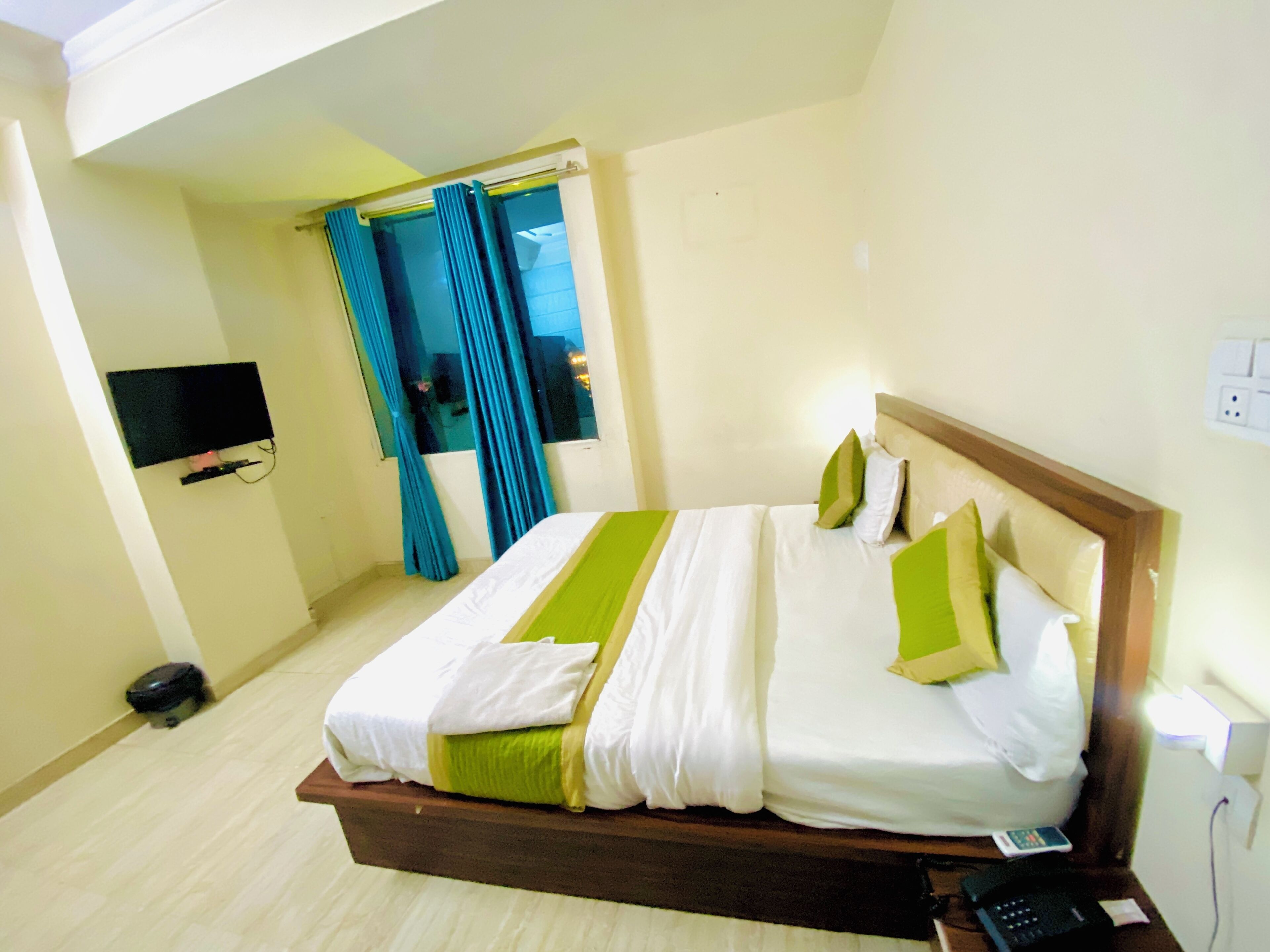 A hotel room with a bed, a TV mounted on the wall, a phone on the bedside table, and a window with curtains.