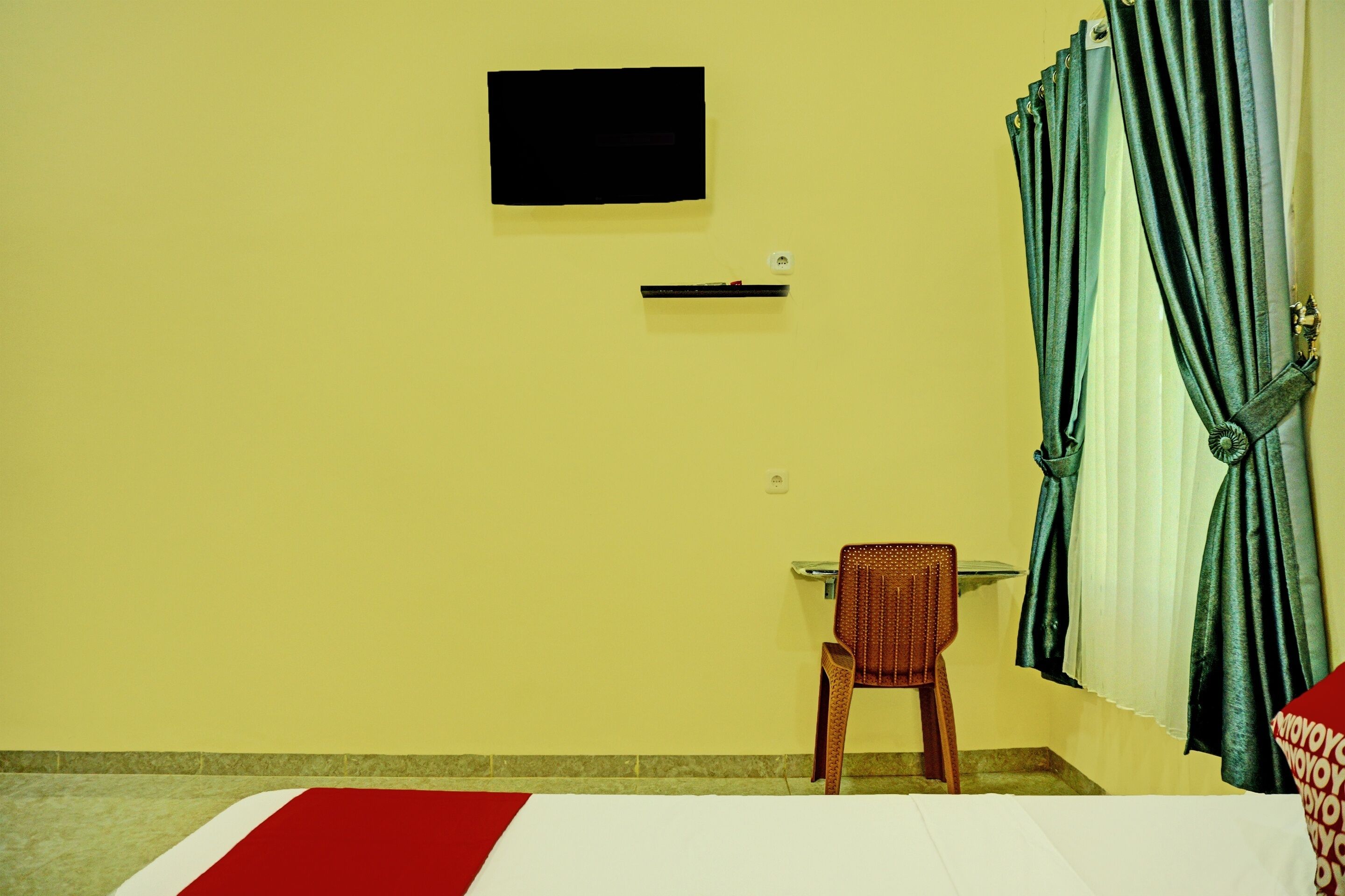 A room with a bed, a chair, a flat-screen TV mounted on the wall, and a window with curtains.