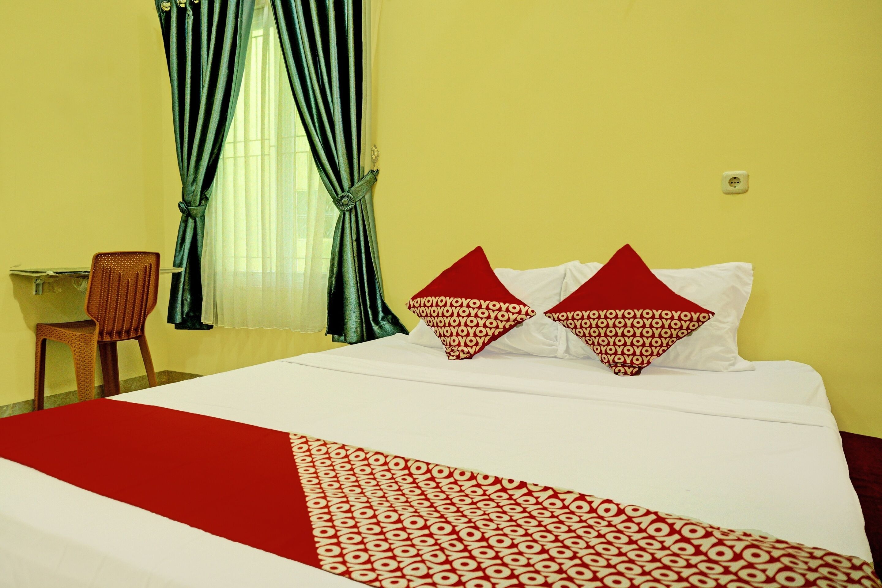 Une chambre d’hôtel avec un lit, deux oreillers à motifs rouges et blancs, une couvre-lit rouge et blanche, une chaise et une fenêtre avec des rideaux verts.