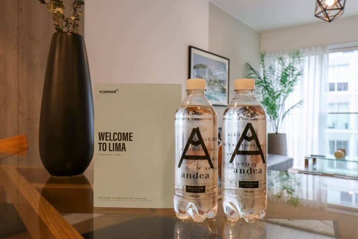 Zwei Wasserflaschen mit den Aufschriften 'A' und 'andea' auf einem Tisch, im Hintergrund ein Schild mit der Aufschrift 'WILLKOMMEN IN LIMA'.