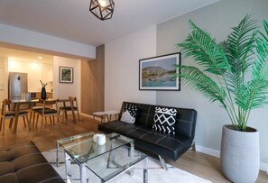 Una sala moderna con un sofá de cuero negro, una mesa de centro de vidrio, un comedor con muebles de madera y una planta grande en maceta.