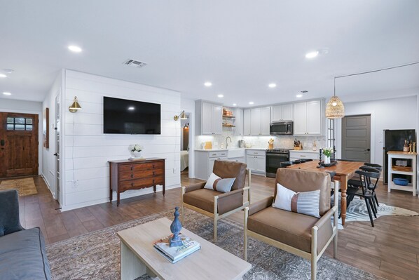 A modern kitchen with white cabinets, a built-in oven, and a microwave. There is a dining area with a wooden table and chairs, and a living room with a sofa and a coffee table. - Ms Dot’s 290 Wine Trail Farmhouse-Downtown- Fire Pit - Covered Patio (Johnson City)
