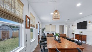 A spacious dining area with a wooden table and chairs, a hanging light fixture, and a view of the backyard. - Ms Dot’s 290 Wine Trail Farmhouse-Downtown- Fire Pit - Covered Patio (Johnson City)