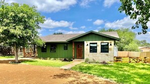 A single-story house with a green exterior, a front porch, and a small stone structure. - Ms Dot’s 290 Wine Trail Farmhouse-Downtown- Fire Pit - Covered Patio (Johnson City)