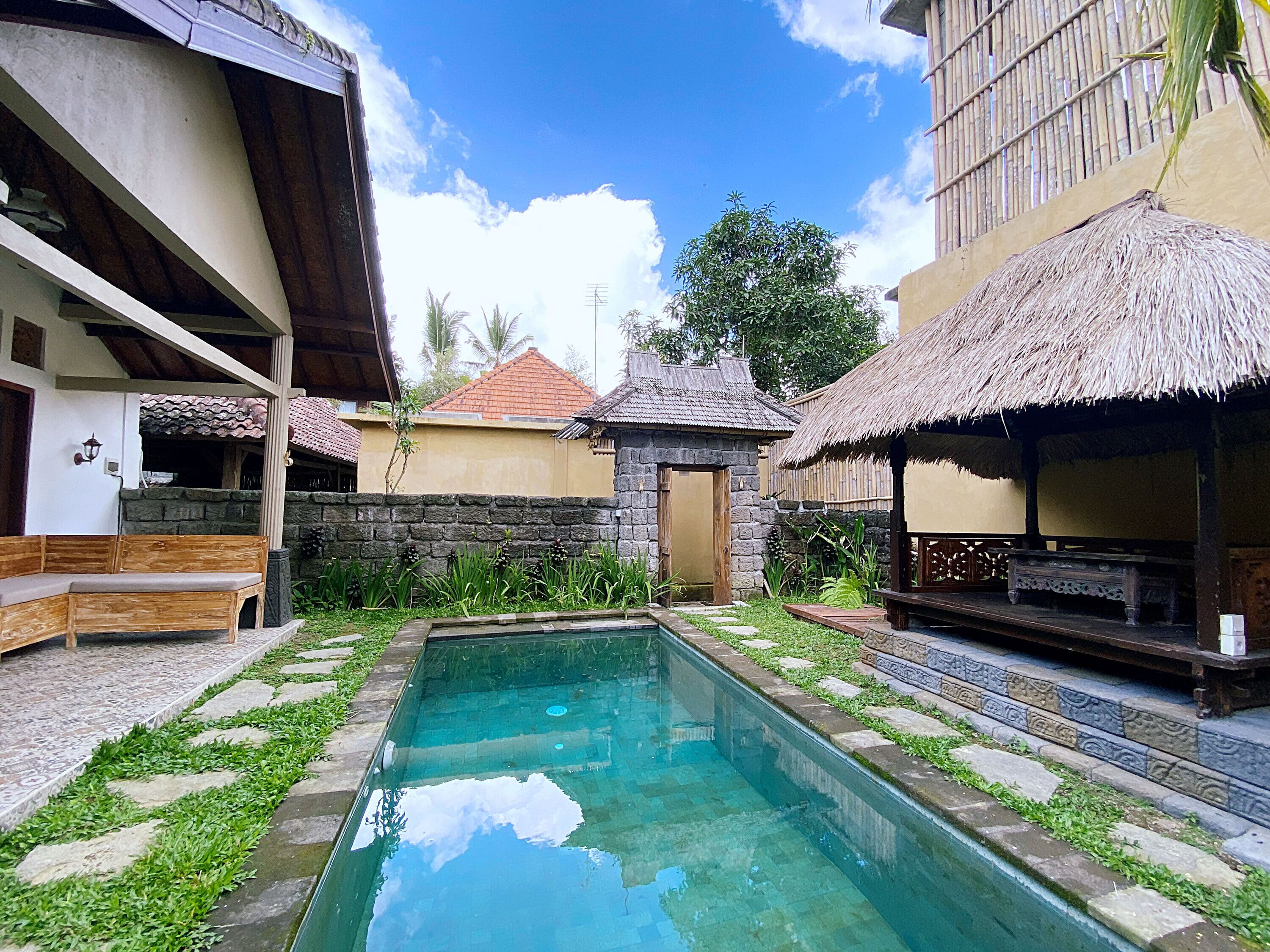 A swimming pool surrounded by stone walls and thatched-roof structures, with a bench and a small garden area.
